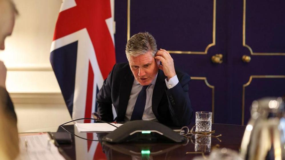 UK Prime Minister Sir Keir Starmer talking on a loudspeaker phone while sitting at a desk with a Union Jack flag situated behind him