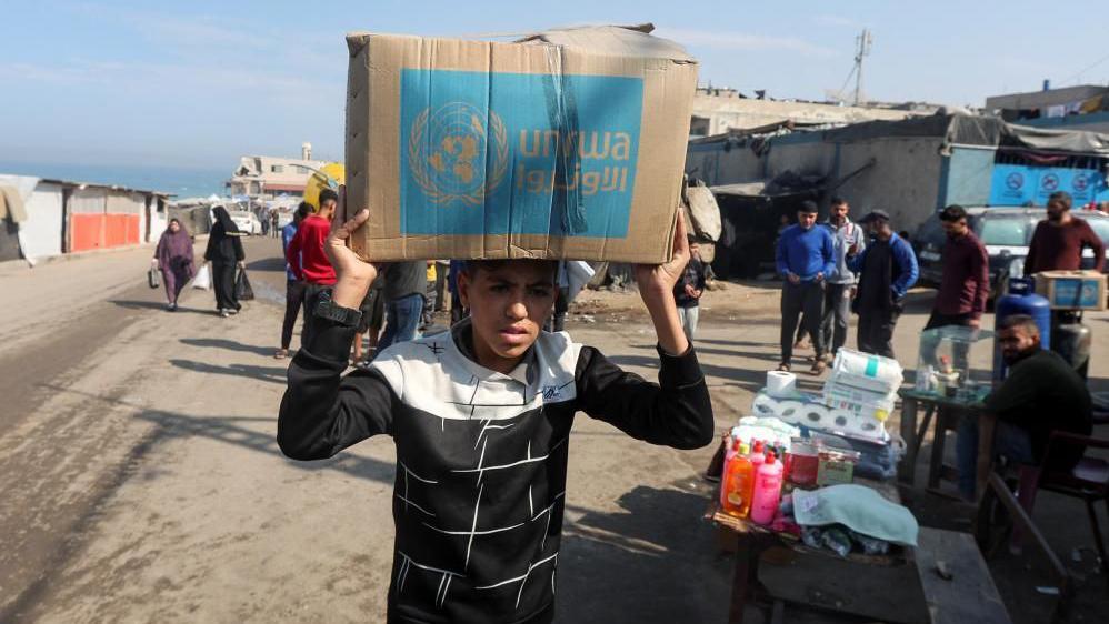 A teenage boy carries a cardboard box with the words "Unrwa" on them as dozens of people stand in the background near some toiletry supplies