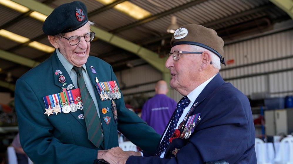 Two World War Two veterans stand together inside an aircraft hangar. Paul Terry, 101, wears a green blazer covered in medals and Ivan Staines, 99, wears a suit jacket and beret with medals attached. Tables and people can be seen in the background.