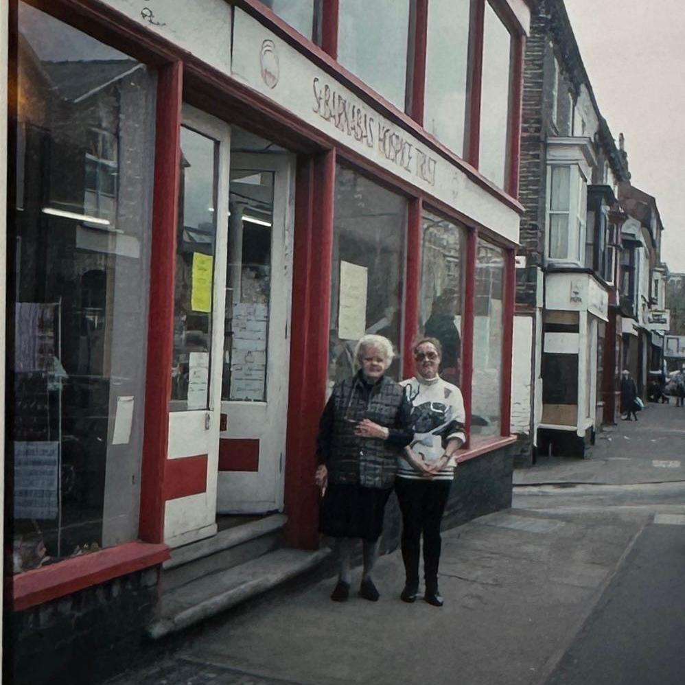 The image shows the front of a red shop named St Barnabas Hospice Trust. The outside has a red frame, with a white door, and there are large glass windows on either side. Above the door, there is a sign with the shop’s name. Two women are standing outside the entrance, holding onto each other’s arm. Other buildings and shops can be seen down the street.
