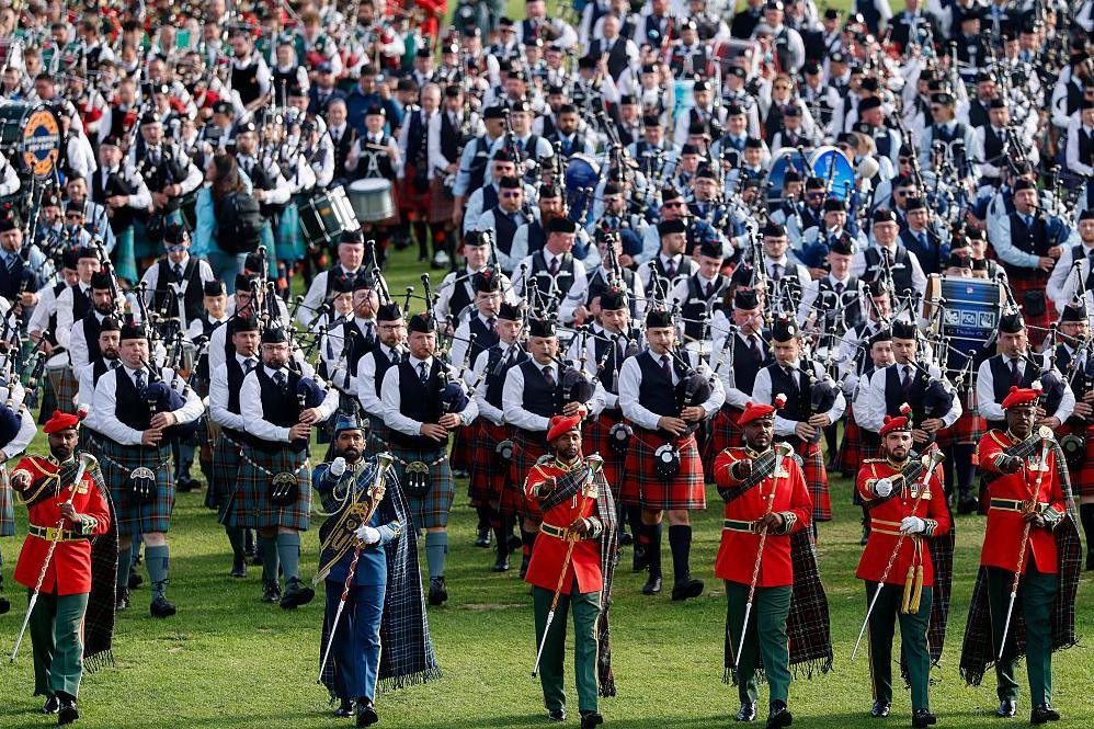 A large gathering of drummers and pipers parading on a field. They are all in traditional outfits and are being led by marching leaders. 