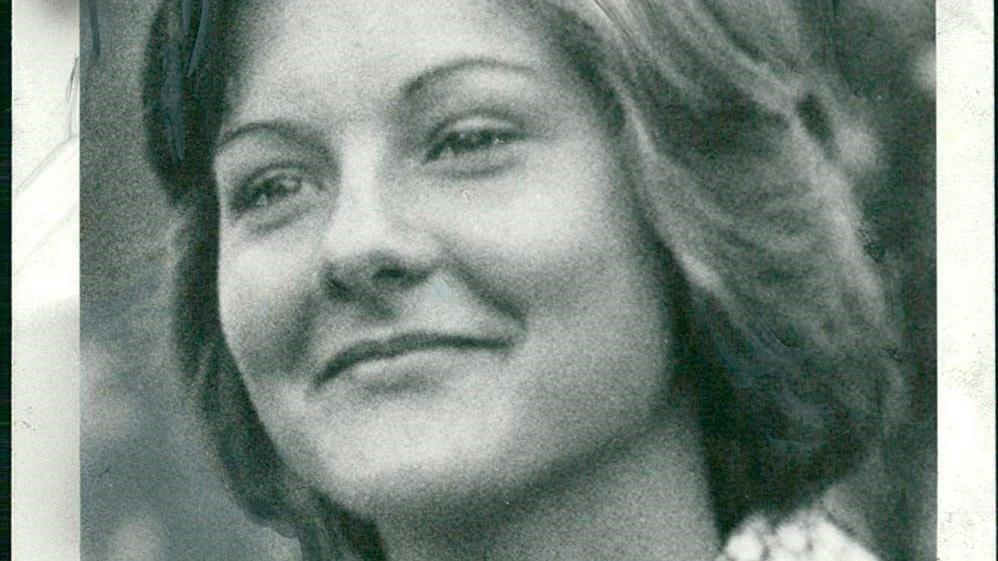 A black and white photo of a smiling young woman looking away from the camera. She is wearing a 1970s-style shirt with a geometric pattern and big wing collars.
