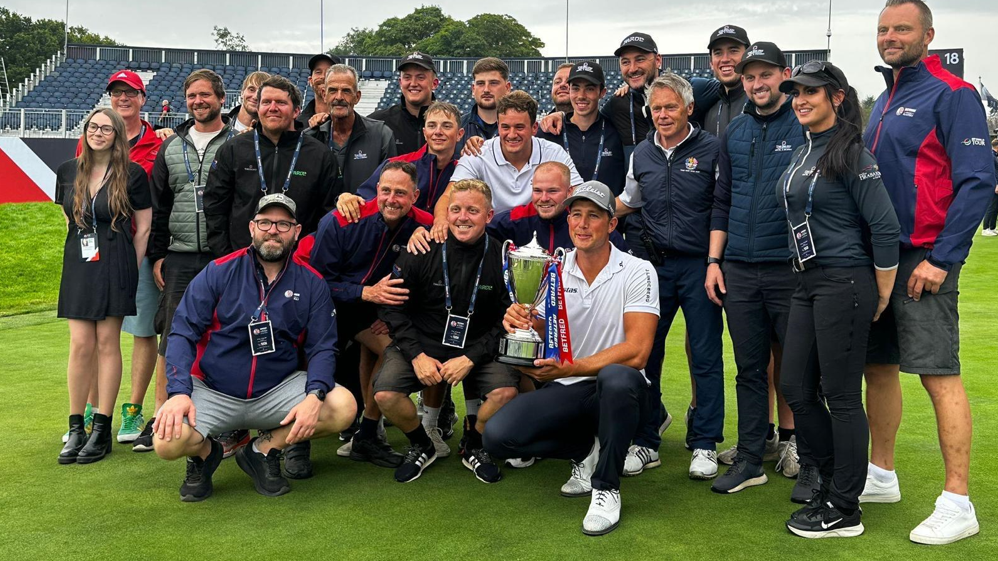 British Masters golfers gather on The Belfry's course with Anna Nilsson. There are more than 20 people in the photo with one man holding a trophy.