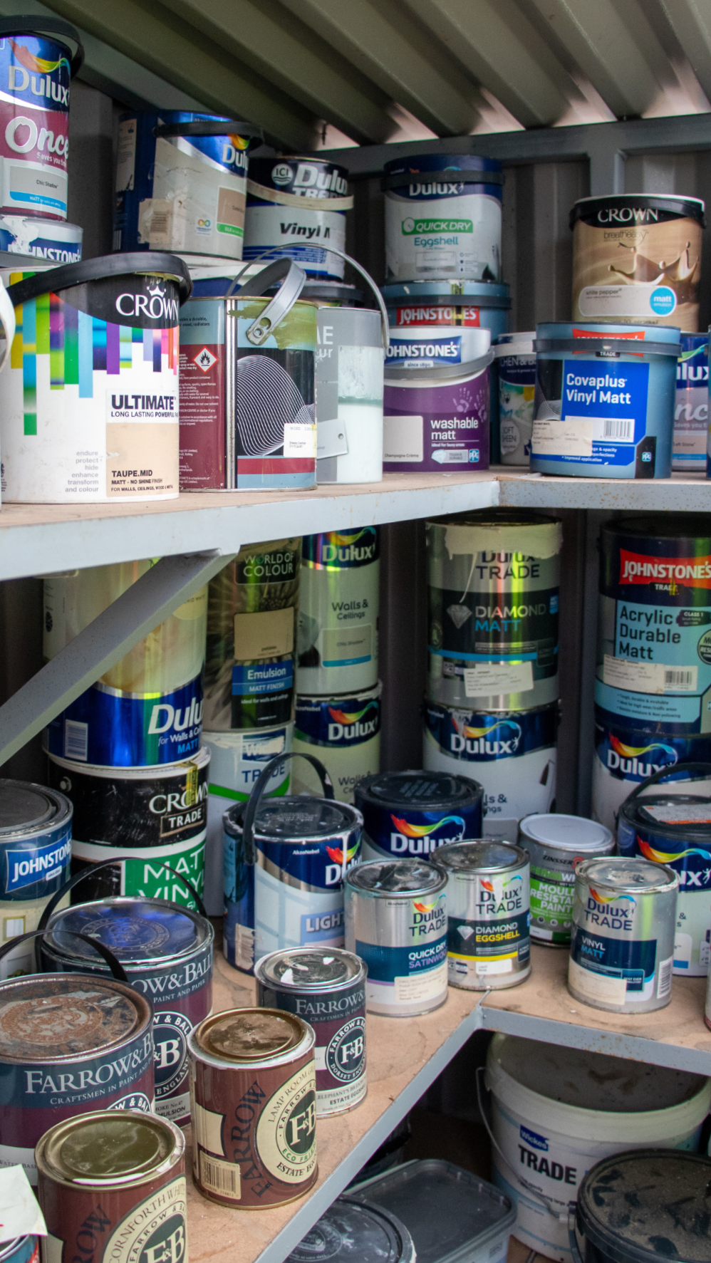 Lots of tins of paint of different sizes and brands on shelves inside a shed.