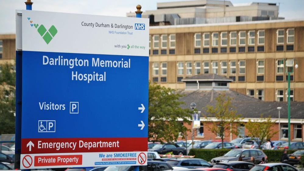 A blue sign outside the Darlington Memorial Hospital. On the top of the sign is a white box which has the logo for the County Durham and Darlington NHS Foundation Trust. In a blue box there are arrows pointing to the car park. A red box below that points to the Emergency Department. Behind the sign is a large, concrete building with many windows and a car park.