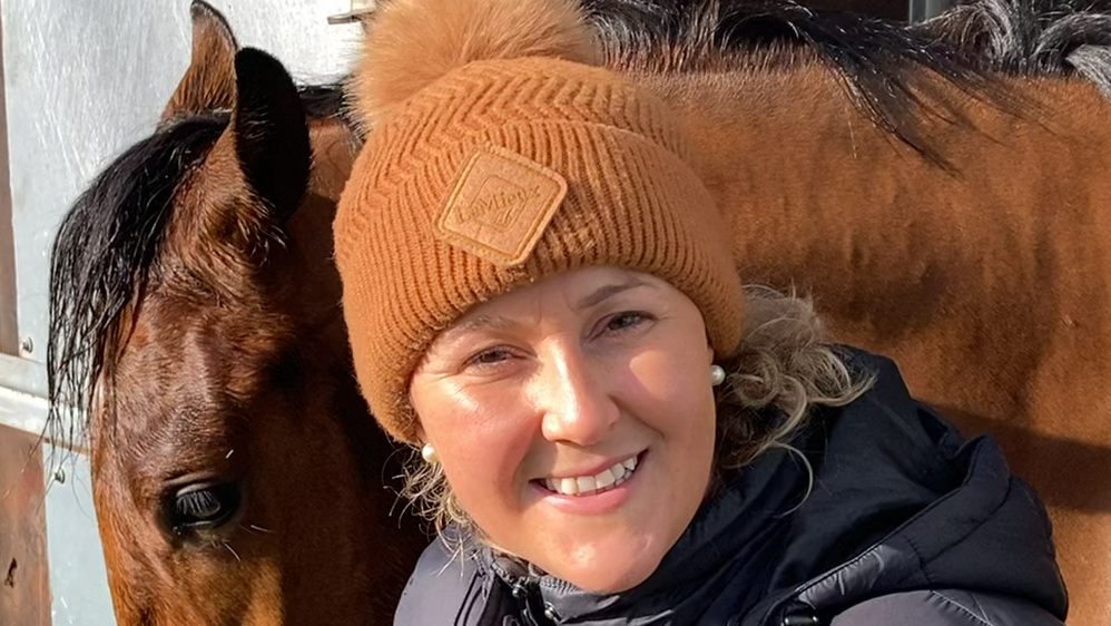 Amy Murphy smiles at the camera with a bay coloured horse with its head hanging out of its stable door. She wears an orange coloured beanie hat with some of her blonde hair hanging out the bottom of it. She also wears a black coat.