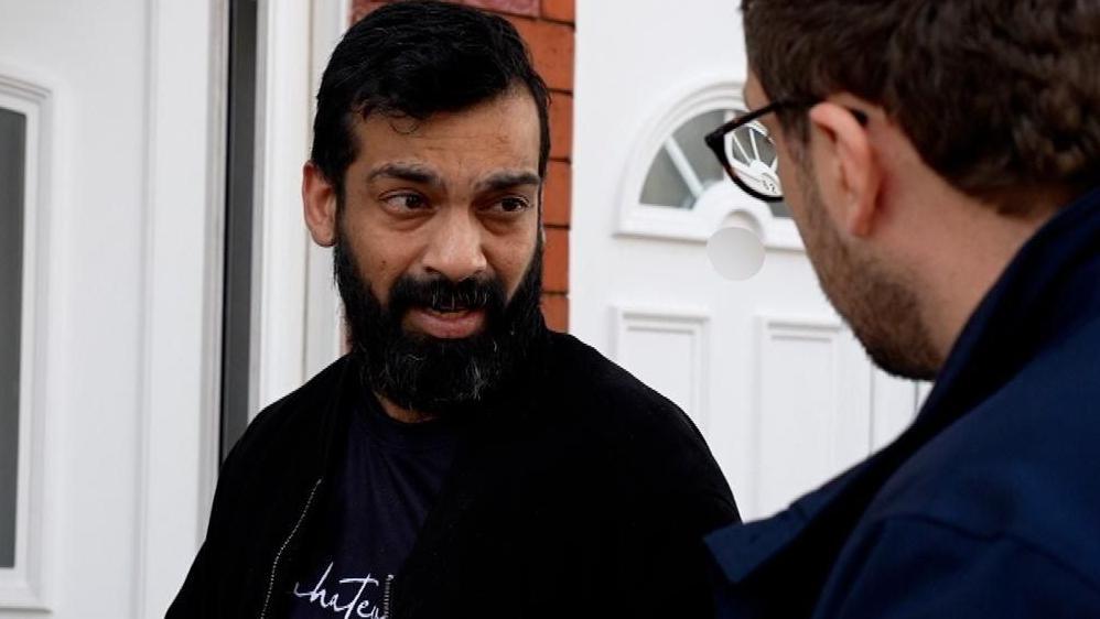 Mr Prabhakar, who has a beard and is wearing a black jacket, stands outside a while front door. With his back to the camera, a BBC journalist questions Mr Prabhakar.
