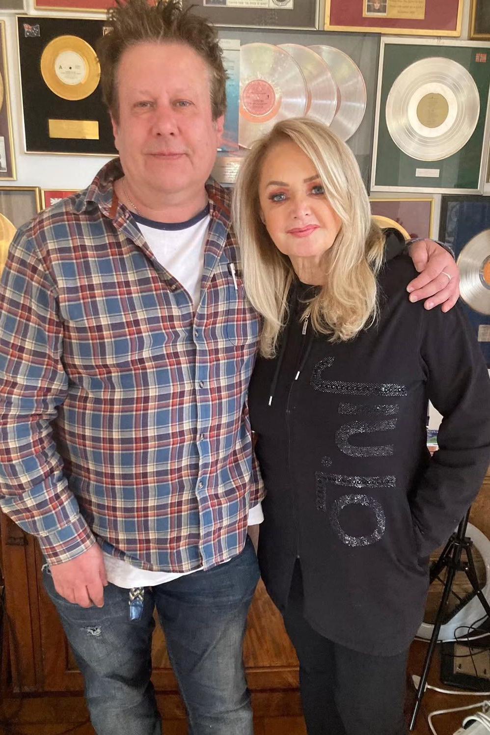 A man with spiky brown hair wearing a red and blue check shirt and jeans, with his arm around a glamorous blonde woman wearing all black. Behind them is a wall with lots of framed records on it.