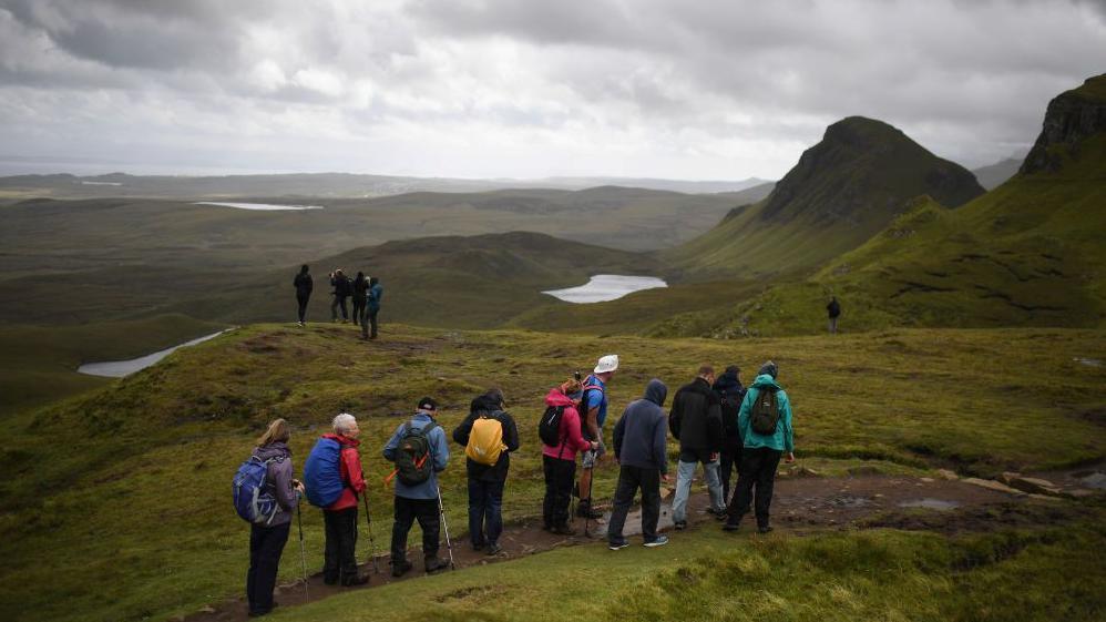 Tourists dressed in outdoor clothing and carrying rucksacks walk along a path on Skye. A separate group are gathered in the distance. They are taking in a wide expanse of hills and lochs under a grey sky.