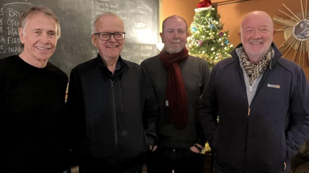 Four men from the band DNA are standing smiling at the camera - there is a Christmas tree and blackboard in the background. 