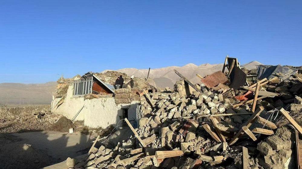 People standing atop a collapsed house, next to a pile of rubble. In the backdrop are mountains.