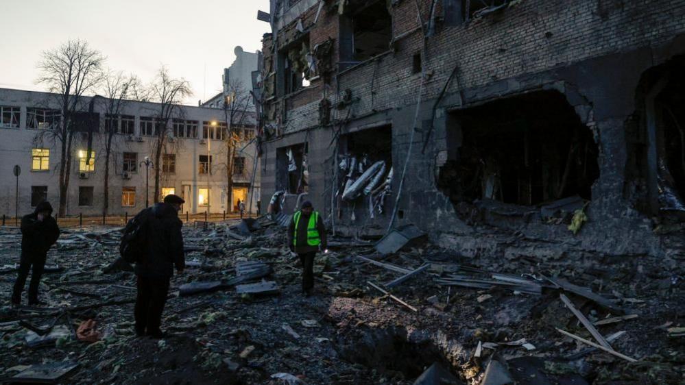 In low light, three men, one in a yellow high-vis vest, stand among the rubble at the base of a badly damaged building