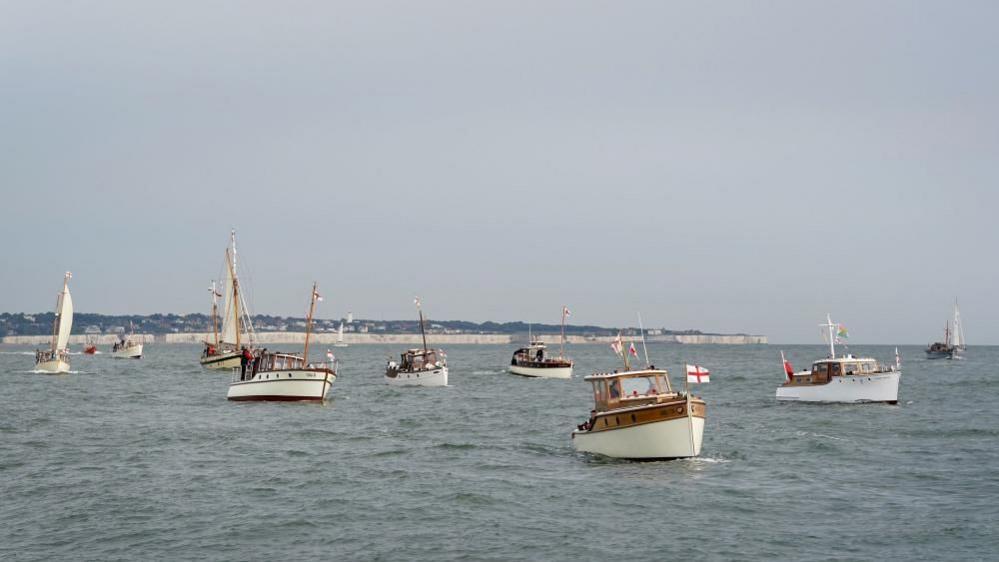 Multiple ships dating back from the 1940s sailing in the sea. Some of the ships are flying St George's flags. The English coast is sat in the background.
