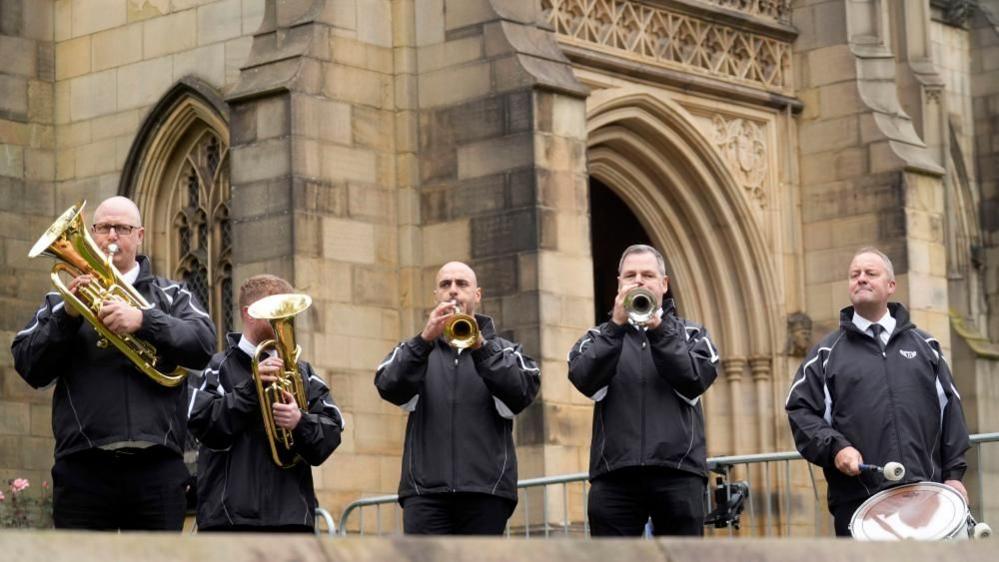 Five men playing wind instruments and a drum stand in front of a grand looking cathedral.