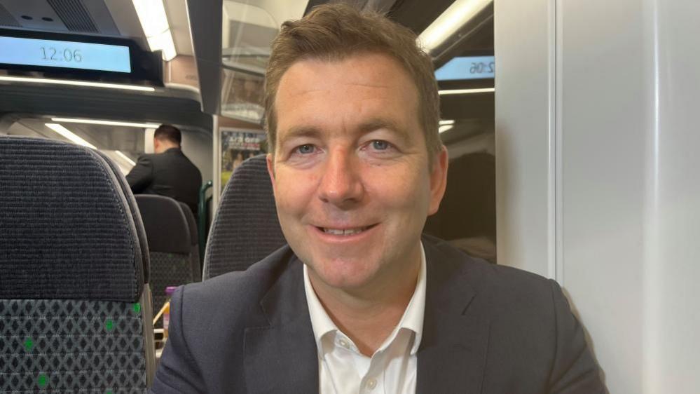 Ian McConnell, sitting in a train carriage, smiling and looking straight at the camera, wearing a white shirt and dark suit jacket. A man is behind him, with his back to the camera.