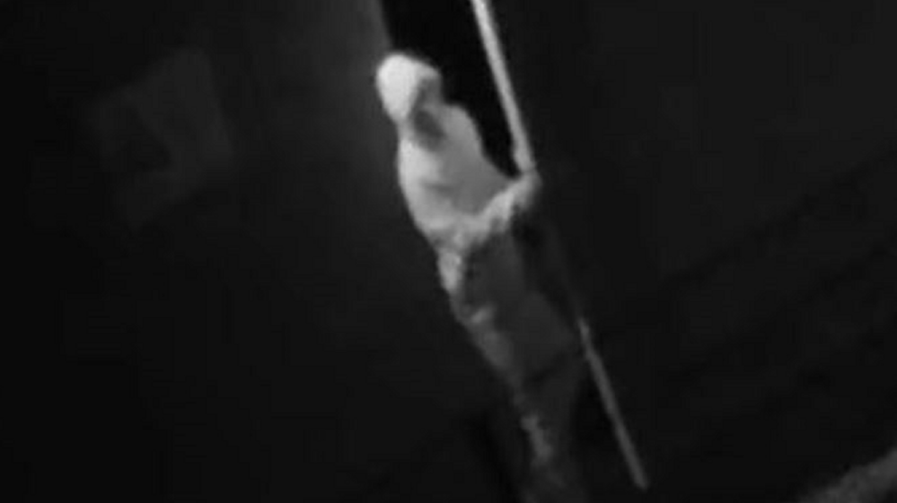 A blurry black-and-white CCTV image shows a hooded man in light clothing opening a door