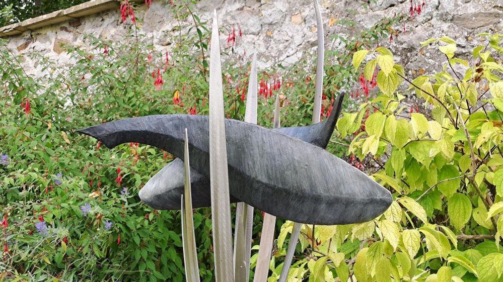 A sculpture in a garden of two simply stylised fish or whale-type metal shapes horizontally weaved in between silver metal pointed sticks coming up vertically which are slightly curved