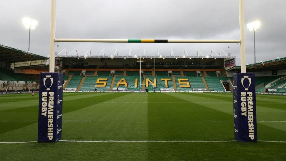 Franklin's Gardens viewed through the rugby posts. Yellow chairs spell out "Saints" on the stand on the far side of the pitch.  The posts are surrounded by Premiership Rugby covers.