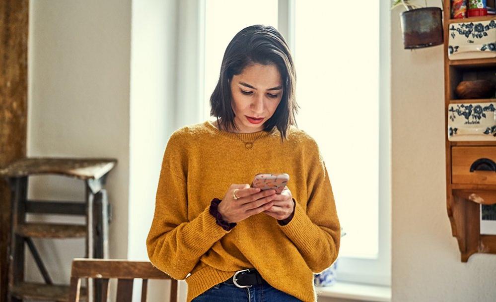 Woman in mustard jumper messaging on a smartphone at home in front of a window