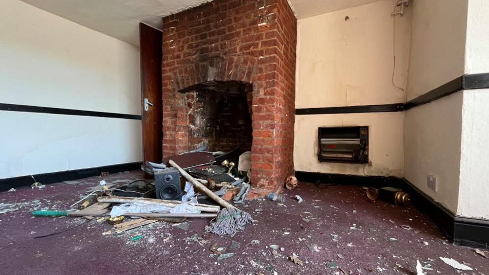 An early 19th Century fire place with alterations in the brickwork, alongside on the wall an electric fan heater. On the floor is a heap of detritus and dirt, on a heavily soiled carpet.