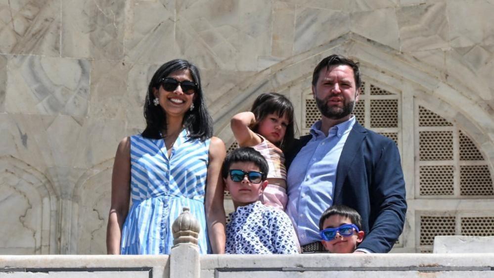 Usha Vance and JD Vance stand with their three kids at the Taj Mahal. JD is holding their daughter. Usha and their two sons wear sunglasses.