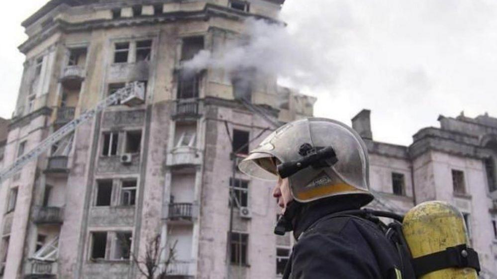 A firefighter walks past a building, fromt which a top-floor window is billowing smoke