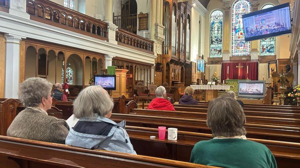 People sitting on brown wooden church pews in St George's Church watching the ceremony on large screens.