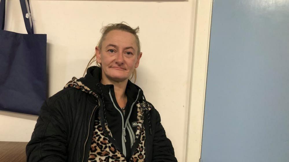 Jade Burtonshaw is sat on a chair in a church hall, she has shaved hair at the sides and it's long on the top and tied in a messy bun. She is wearing a animal print onesie and multiple layers. 