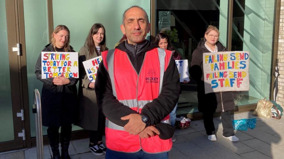 A man, wearing a red vest, stands in front of a line of women holding colourful signs . One reads: "Striking today for a better tomorrow". 