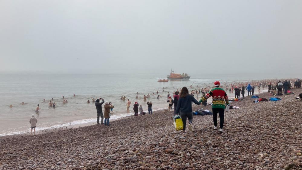 People on a shingle beach look on as dozens of people take to the sea - watched over by a RNLI boat