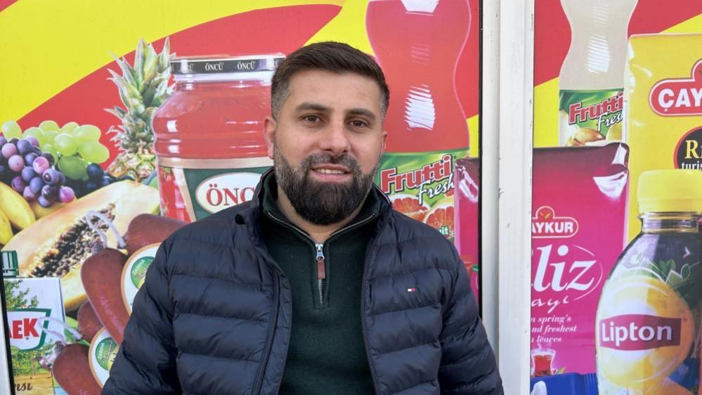 Shahrim Ali with short dark hair and medium-length beard, wearing a dark top and blue gilet, standing in front of a shop window which has illustrations of various products.