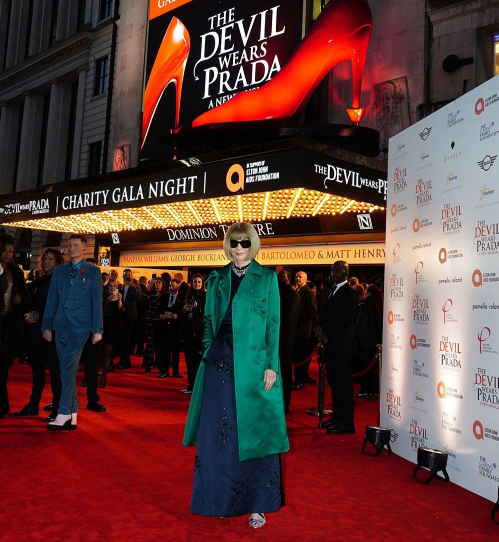 Anna Wintour standing outside the Dominion Theatre in London which displays a gala night sign reading 'The Devil Wears Prada' with two red high-heeled shoes.