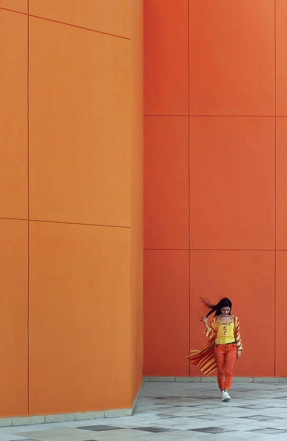 A young woman in bright orange and yellow clothing posing beside a similarly orange wall