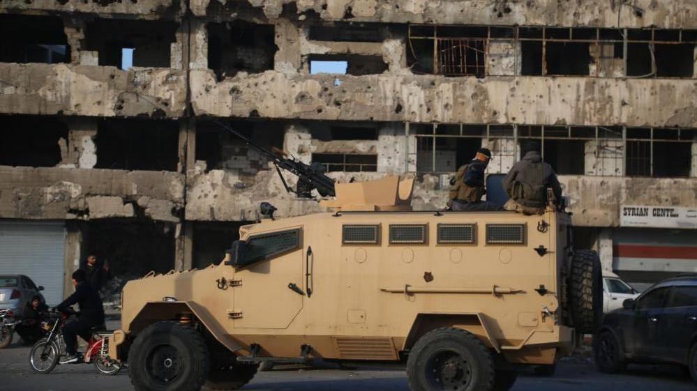 sand-coloured armoured car in front of a ruined building in Homs