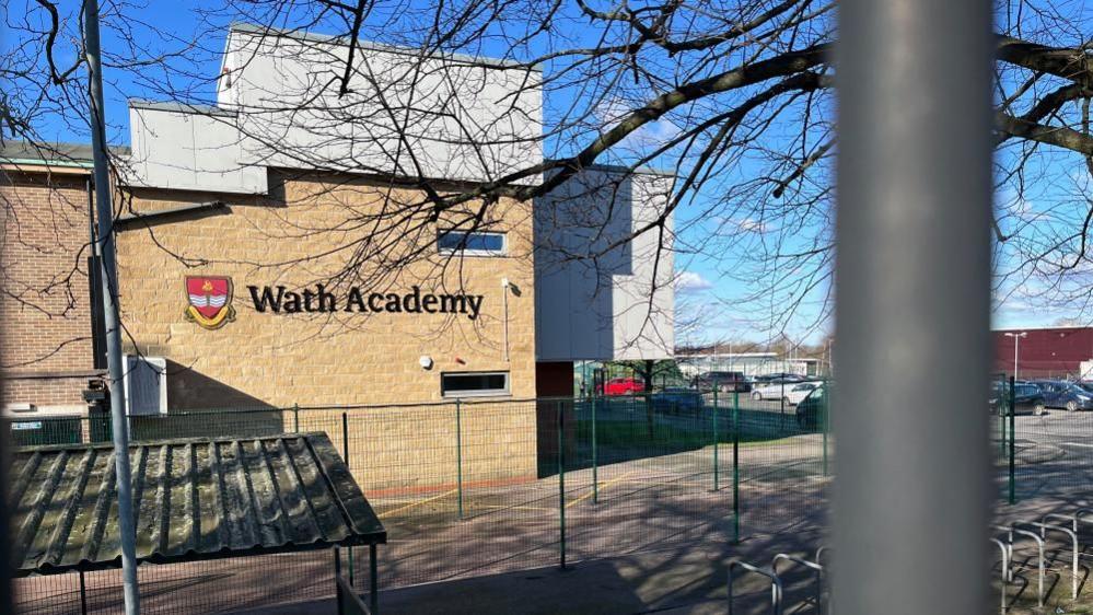 A photo of a sandstone building with "Wath Academy" on the side. There is a car park to the right of the building and what looks like a bike shelter to the foreground.