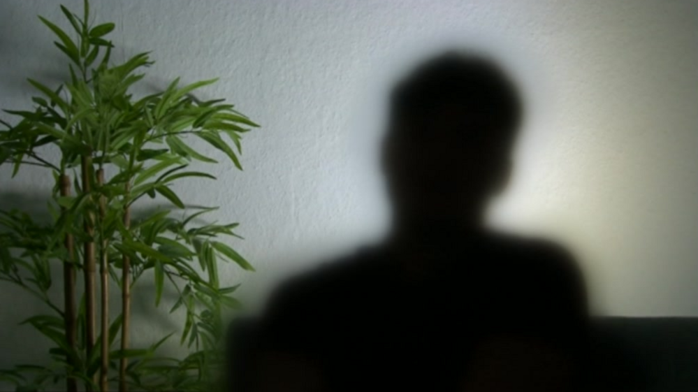 A blurred silhouette image of the victim, showing their head and shoulders against a white wall. There is also a potted plant on their left