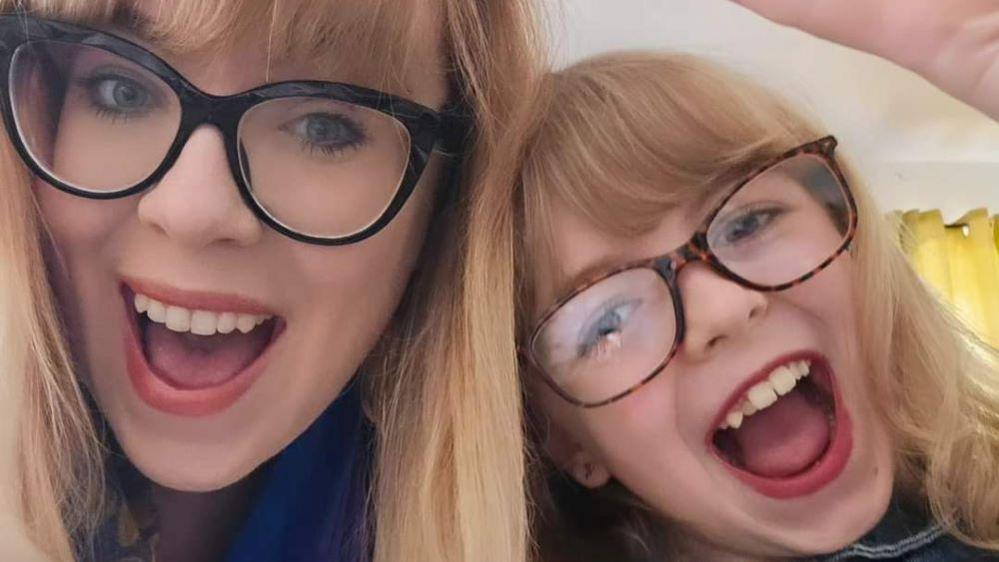 Katrina and her daughter Rosalie. Both have long blonde hair and are smiling open-mouthed at the camera. Katrina has black-rimmed glasses and Rosalie has brown-rimmed glasses.