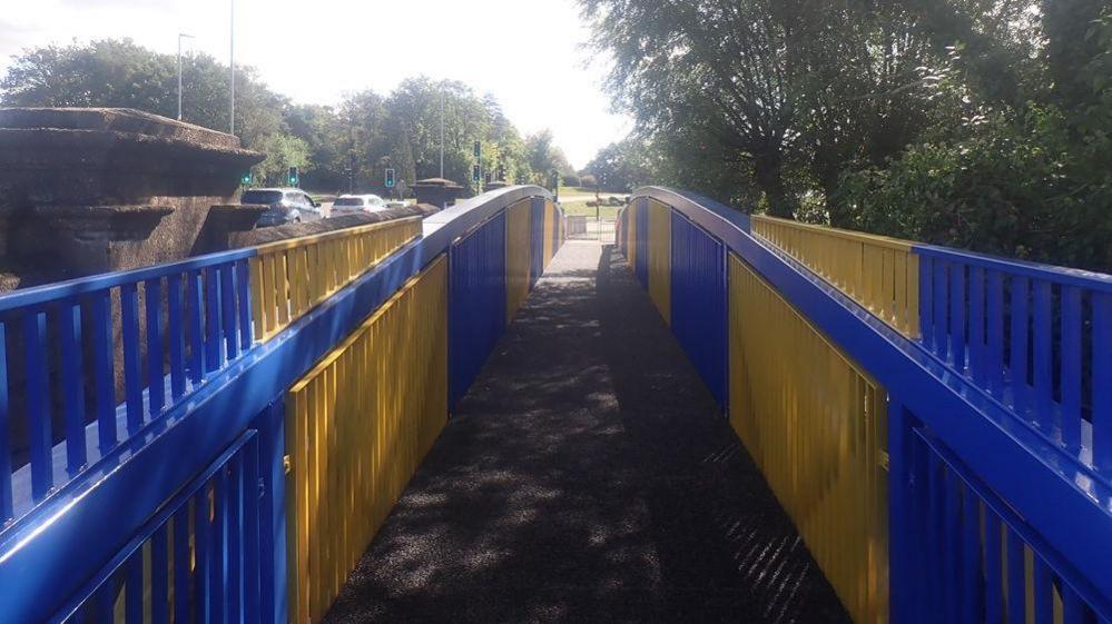 A blue and yellow painted metal footbridge that runs parallel to a bridge with cars going over it. In the distance are traffic lights and a large roundabout