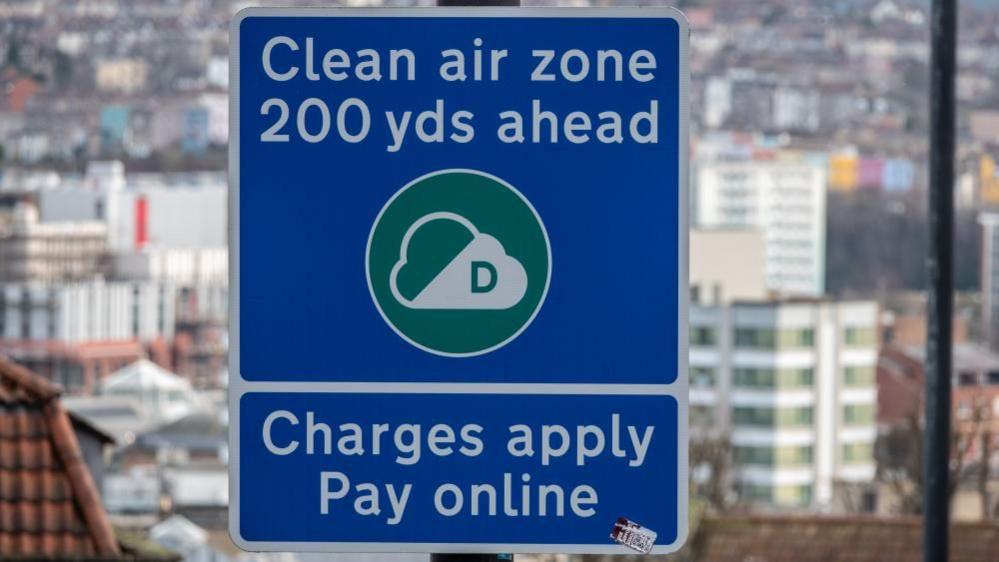 A blue Clean Air Zone sign in front of a Bristol skyline, which reads: 'Clean air zone 200 yds ahead. Charges apply, pay online'