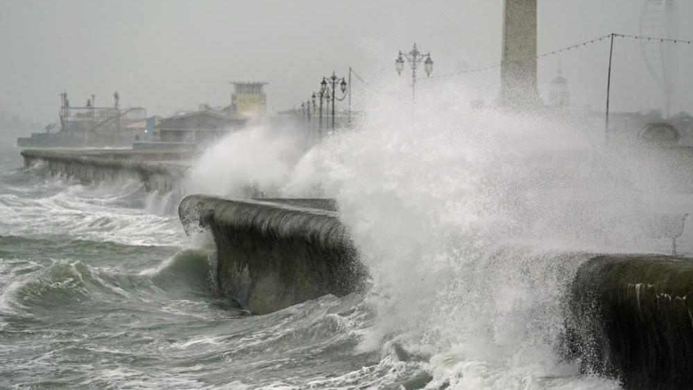 Waves crash against a sea wall, spilling onto the land. There is lots of spray from the waves. The sky is grey and on land there are street lights and buildings visible in the background.