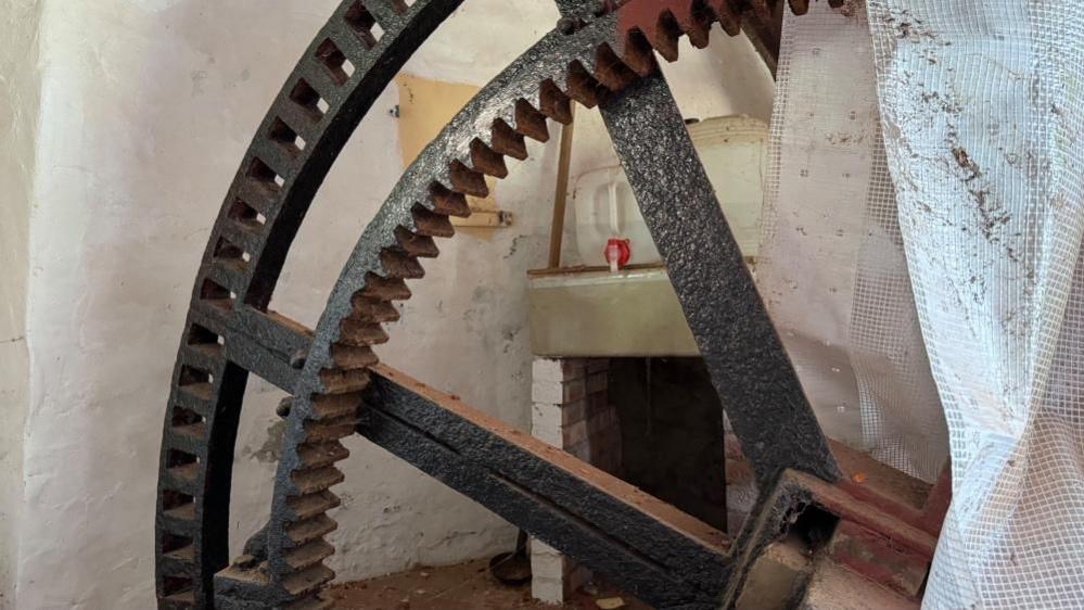 A very large cast iron gear wheel, partially covered with reinforced plastic sheeting. The wheel features a traditional gear wheel on an inner ring, with holes in the outer ring that would hold oak teeth that would shear sacrificially in abnormal conditions to protect the structure from severe damage.