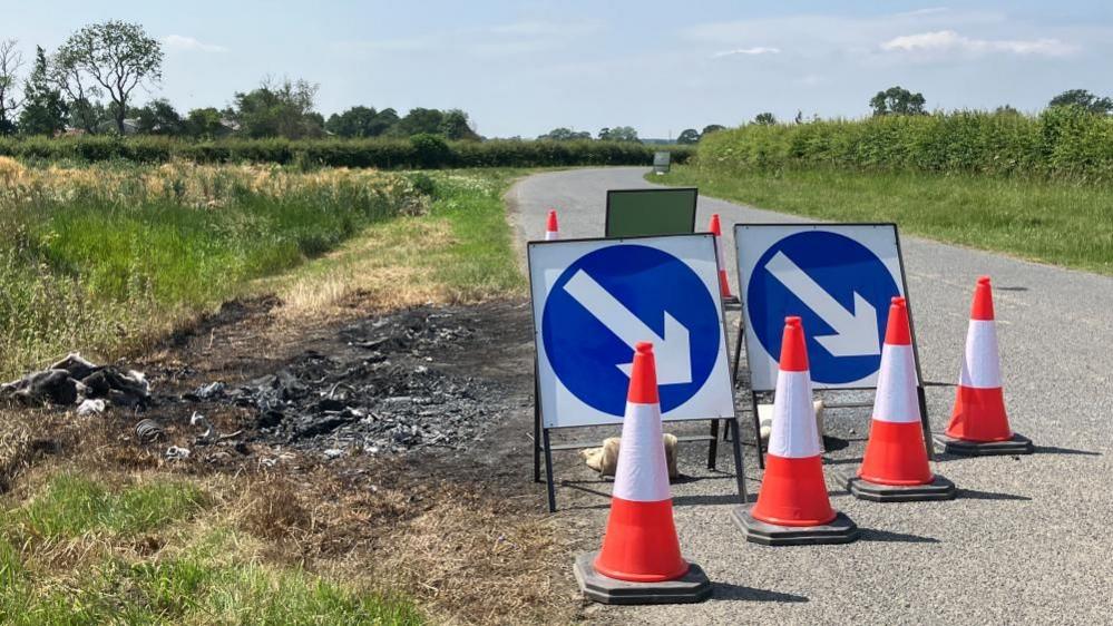 A series of cones and road signs send drivers around a scorched patch of earth at the side of a country road