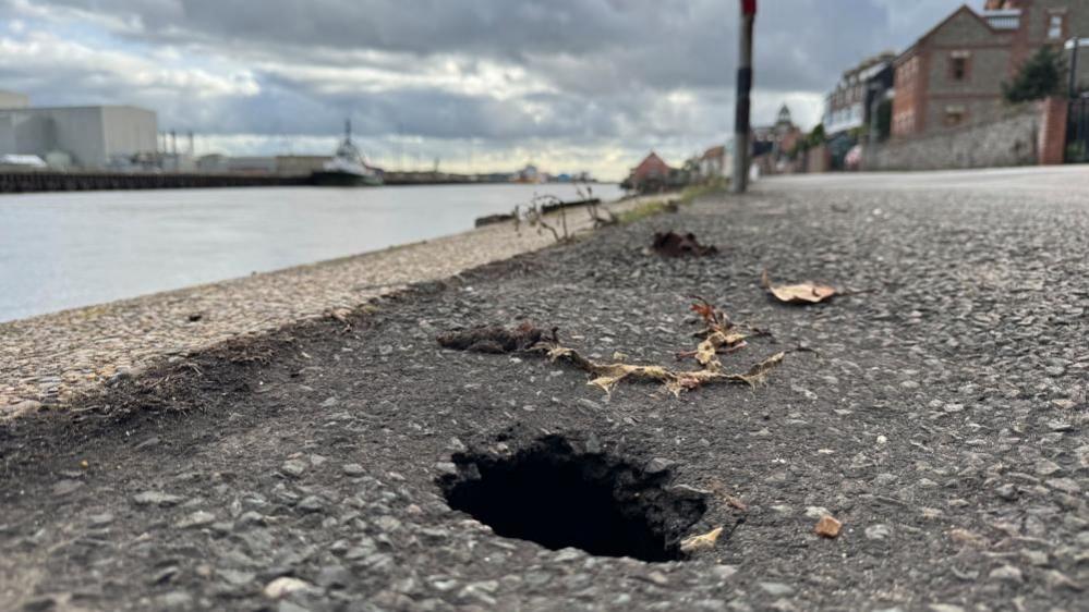 A road surface, with a hole visible in the near-ground. Beyond is the River Yare and ships and Great Yarmouth Power Station. Houses are visible on the rightmost far distance.
