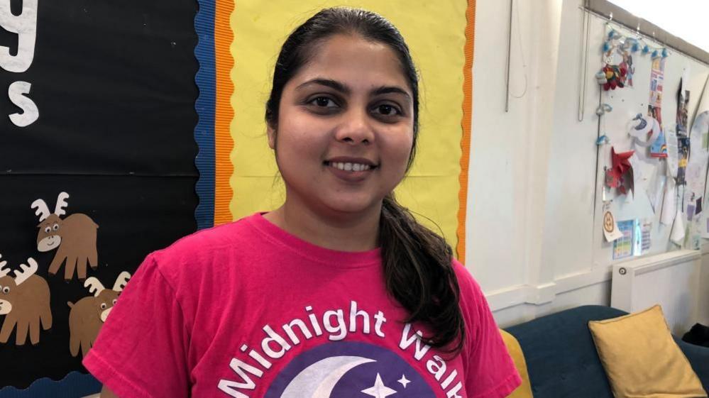 A woman is smiling at the camera she has long dark hair and s wearing a pink t-shirt. 