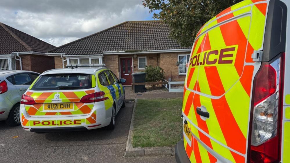 A police van and a police car parked outside the home of Darren Mahoney. The back doors of the van, bearing high visibility chevrons in orange and yellow can be seen to the right of the image; the car, fully marked, is parked front-end towards Mahoney's house, a semi-detached bungalow of 1990s construction with a hipped, tiled roof. A lawn and tree are seen to the centre/right of the image.