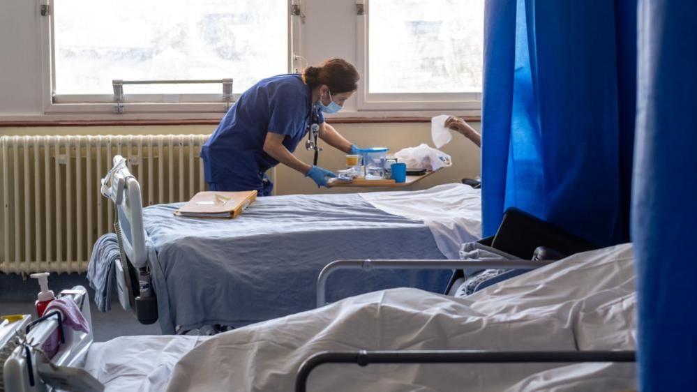 Inside a hospital ward, two patients can be seen lying in hospital beds. Their faces can't be seen as they are behind the divider curtains. A nurse wearing a face mask speaks to one of them.