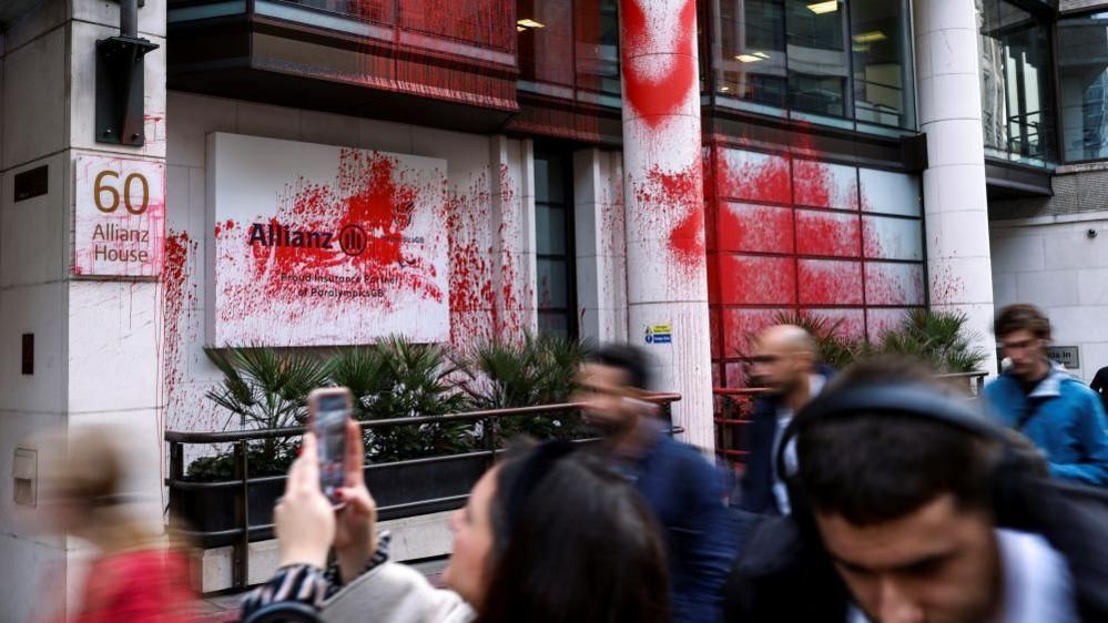 White-fronted office building in London with pillars between the windows. Much red paint is visible on the frontage of the building - including on the Allianz sign.  People are walking past and one is taking a picture on a mobile phone.