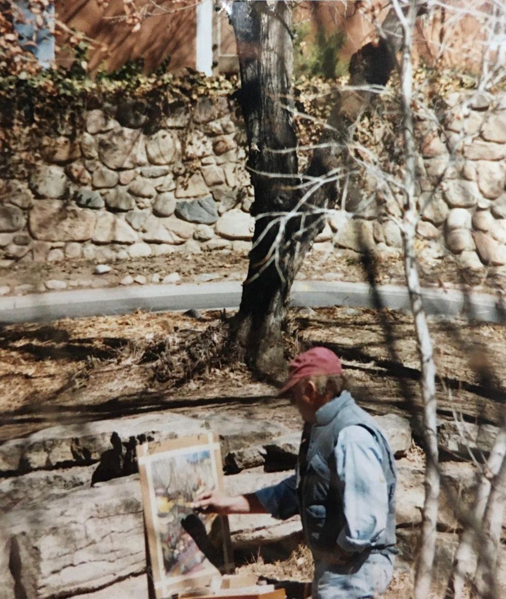 A man wearing a red cap and blue shirt is seen painting in a landscape