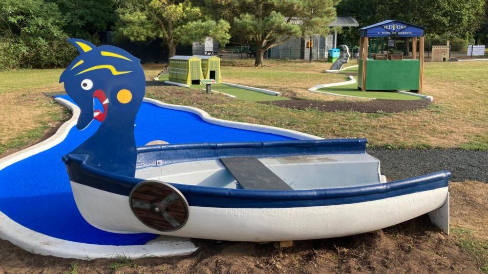 A blue and white boat on a crazy golf course in Bedford, with replicas of the Cardington hangars and a market stall in the background