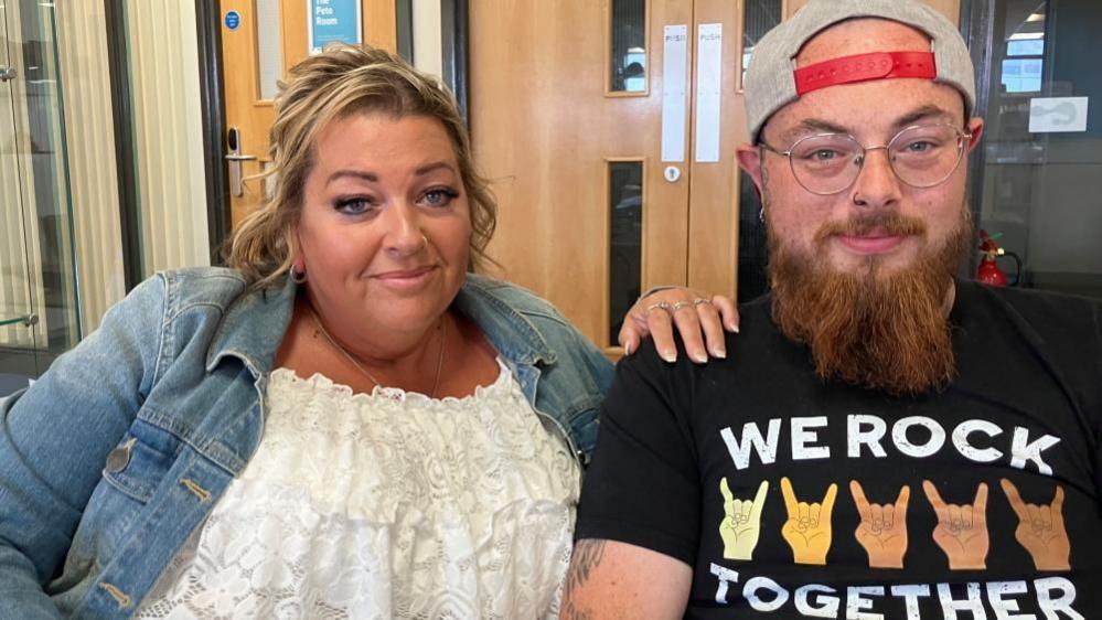 Connor Parker is wearing a sand-coloured baseball cap with red strap backwards. He is wearing a black T-shirt with "we rock" on the front. He is smiling at the camera, wearing glasses and he has a nose ring. He is sitting next to his mother who is wearing a white dress and blue jeans jacket.